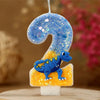 A birthday candle shaped like the number 2, decorated with blue glitter and a small dinosaur figure.