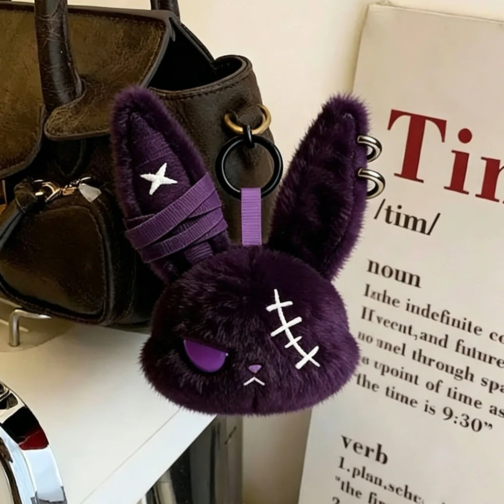 A dark purple gothic bunny plush keychain with a bandage design is displayed next to a black leather bag.