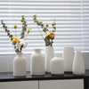 A collection of white ceramic mini flower vases with dried greenery and small yellow and orange flowers set against a white s