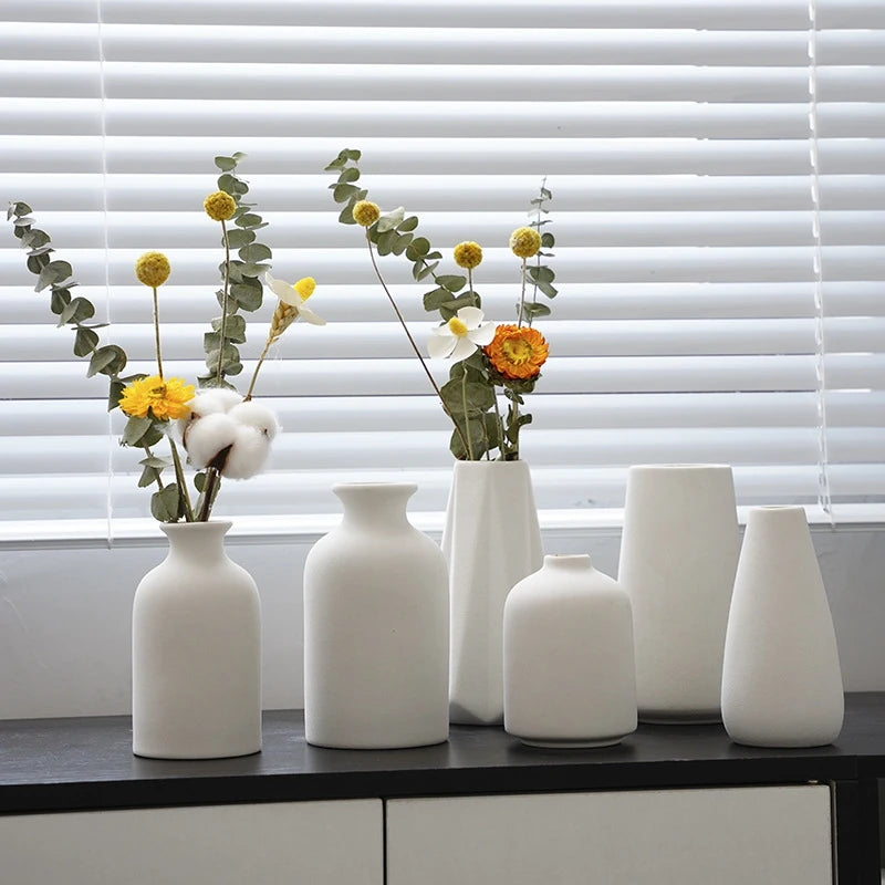 A collection of white ceramic mini flower vases with dried greenery and small yellow and orange flowers set against a white s