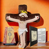 A plush doll depicting Jesus on a cross with a crown and beard, set against a warm orange background next to a Bible.