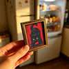 A hand holds a wooden magnetic cat photo frame featuring a black cat silhouette against a red background.