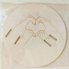 A wooden baby announcement sign featuring a line drawing of two hands forming a heart.
