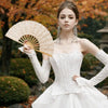 A model wearing a white lace dress holds an open beige hand fan with a tassel pendant in an outdoor setting.