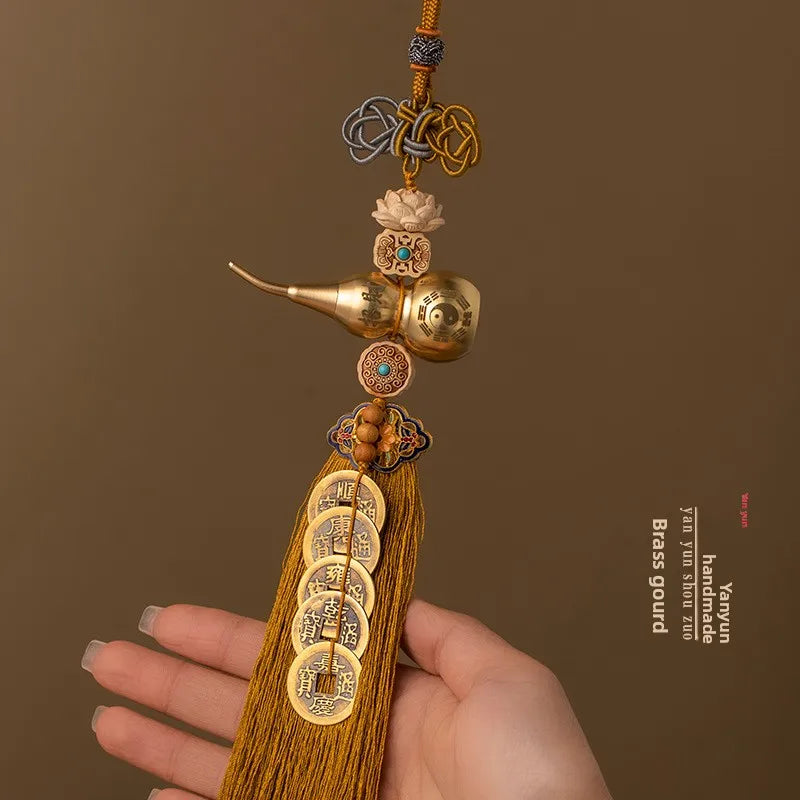 A hand holds a brass gourd and five emperor coins hanging ornament made of gold-toned metal and yellow thread.
