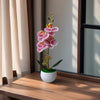 A realistic artificial orchid with pink flowers is potted in a white ceramic pot on a wooden surface.