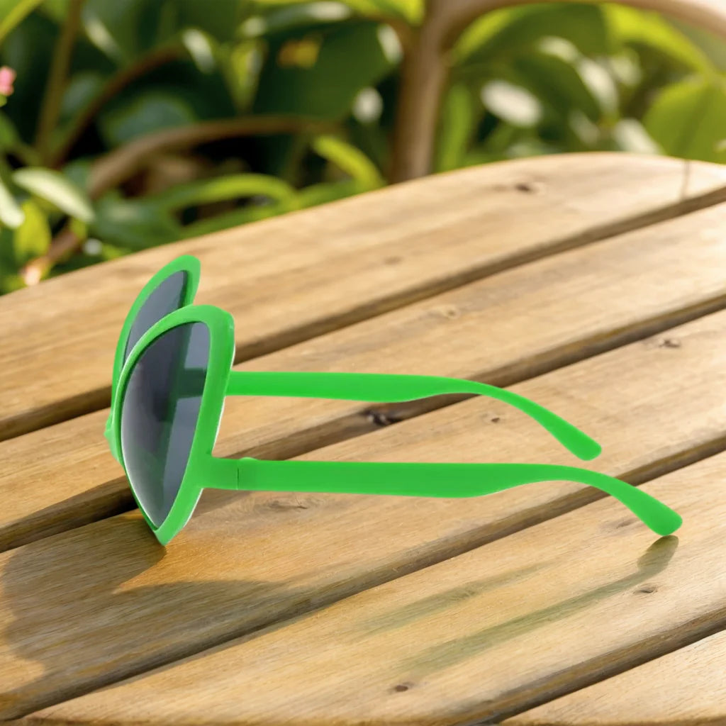 A pair of bright green alien sunglasses resting on a wooden surface with blurred greenery in the background.