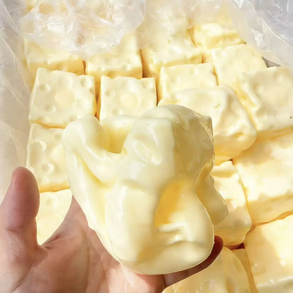 A hand holds a block of yellow cheese squishy stress relief toy surrounded by other similar blocks in clear plastic packaging