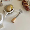 A manual wooden potato and pumpkin masher resting on a white table next to a whole potato and a loaf of bread.