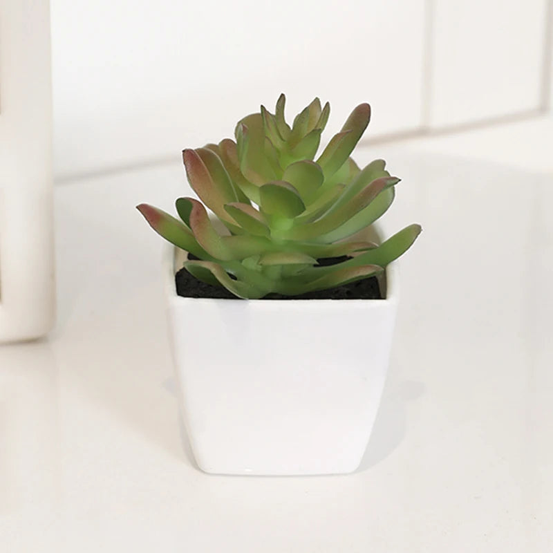 A small, green artificial succulent plant in a white pot sitting on a white surface.