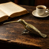 A vintage brass hummingbird statue resting on a dark wooden surface next to a cup and saucer.