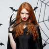 A woman with long red hair wearing black clothing and black skeleton hand gloves against a dark background.