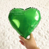 A bright green heart-shaped foil balloon being held by a person on a white textured surface.