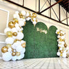 A balloon garland featuring white and gold latex balloons set against a green hedge backdrop.