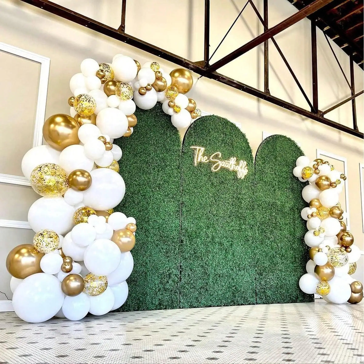 A balloon garland featuring white and gold latex balloons set against a green hedge backdrop.
