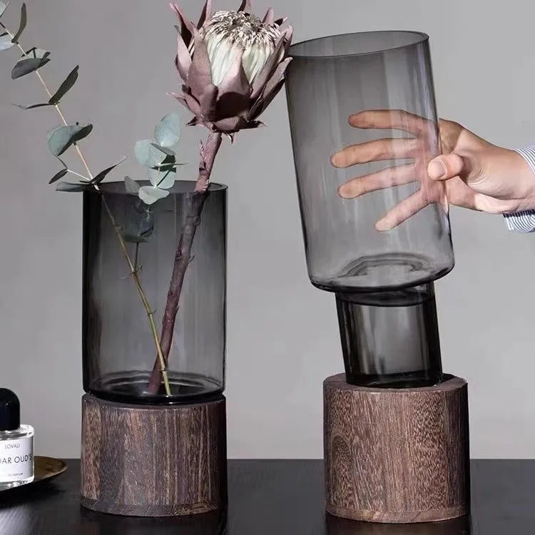 A hand holds a tall, cylindrical glass vase with a wooden base, set against a gray background.
