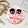 A round button pin featuring a skull and crossbones design with a heart and a cross, set against a pink background.