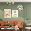 A rustic wooden wall clock with a distressed white face hangs on a sage green wall above a brown sofa.