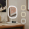 A white vanity mirror with three illuminated light settings: warm, mixed, and cold light, displayed on a wooden surface.