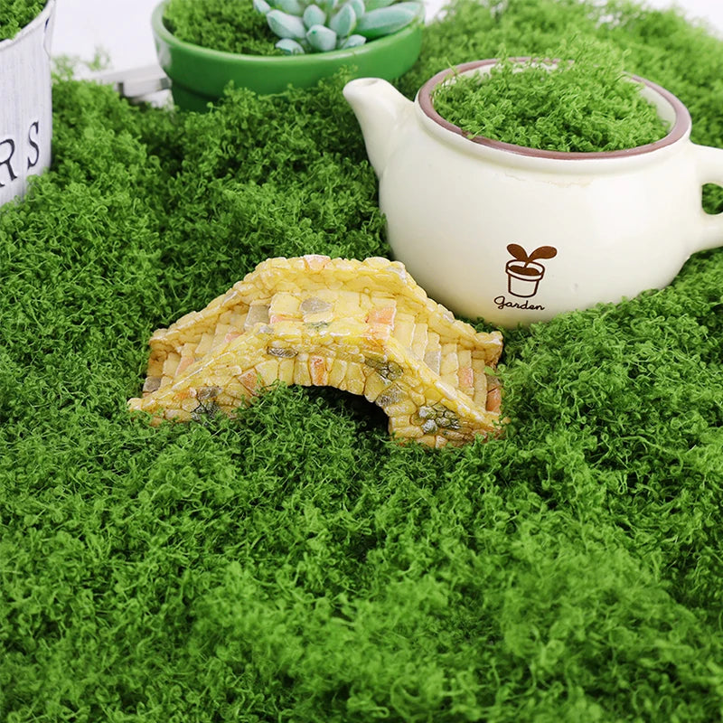 A piece of textured yellow artificial moss rests on bright green faux moss with a white teapot and a succulent plant in the b