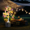 A glowing butterfly birch tree light illuminates a wooden table set outdoors under a white tent.