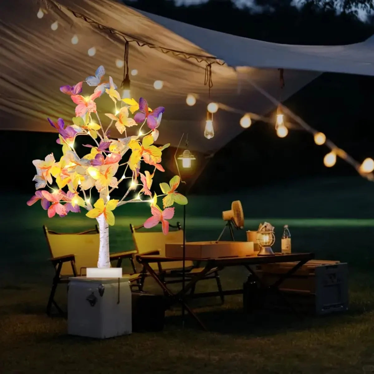 A glowing butterfly birch tree light illuminates a wooden table set outdoors under a white tent.