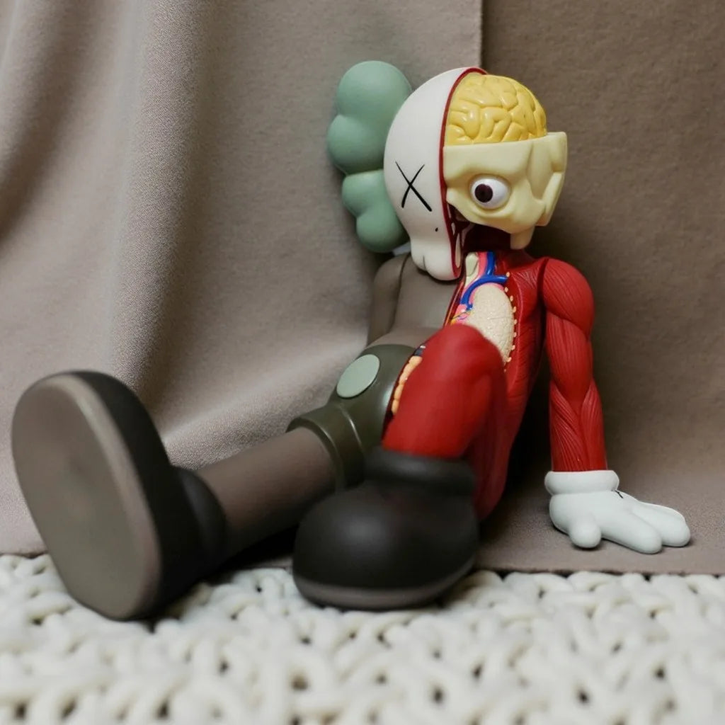 A stylized figurine of a bear with a yellow head, red body, and green ear, sitting on a light fabric surface.
