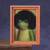 A framed picture of a green frog with black curly hair, displayed in a wooden photo frame.