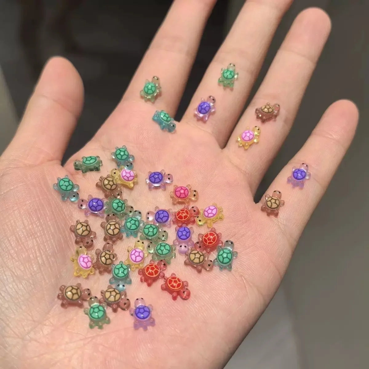 Una mano muestra 50 adornos de uñas de tortugas mini y coloridos esparcidos por los dedos.