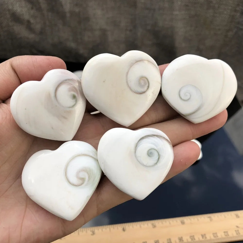 Five white rose quartz heart-shaped stones with swirling patterns are held in a hand above a ruler.