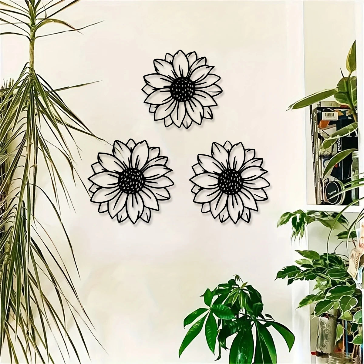 A set of three black metal sunflower wall decor cutouts displayed on a white wall next to a large green plant.