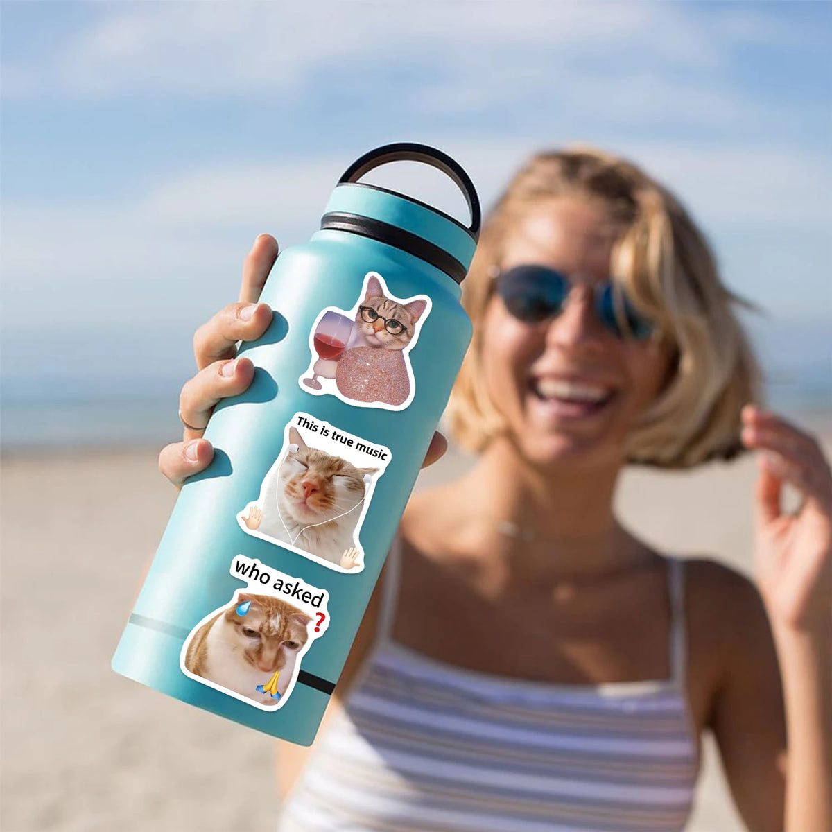 A woman holds a teal insulated water bottle with a cat decal on it outdoors.