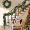 An artificial cedar garland draped along a wooden staircase railing in a bright interior setting.