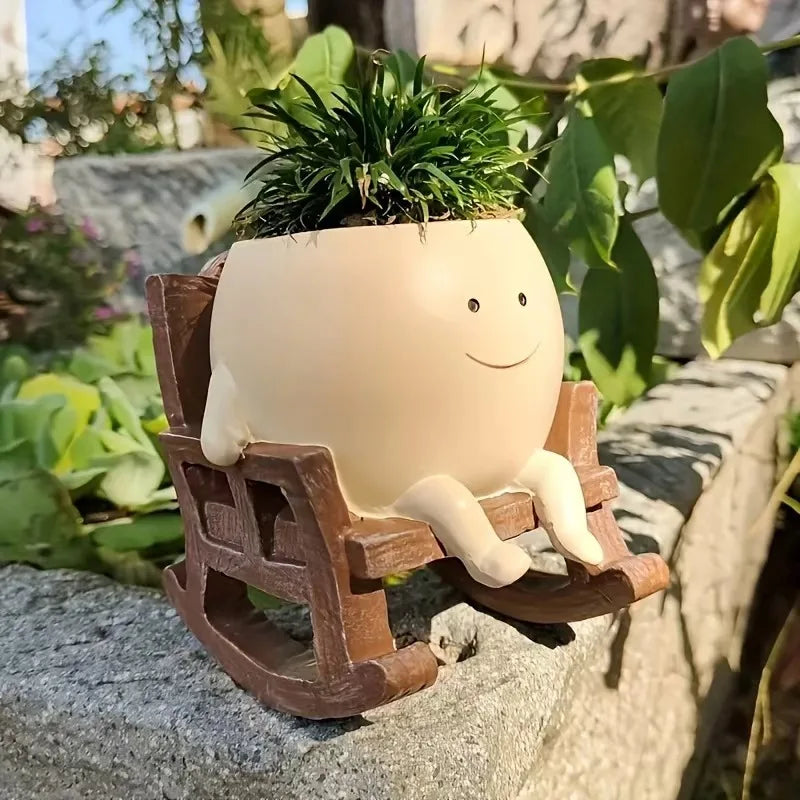 A resin ornament shaped like a smiling face with green foliage, resting on a wooden planter outdoors.