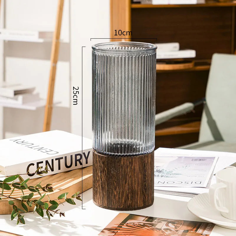 A tall glass vase with a ribbed texture and a dark wooden base is shown on a white surface.