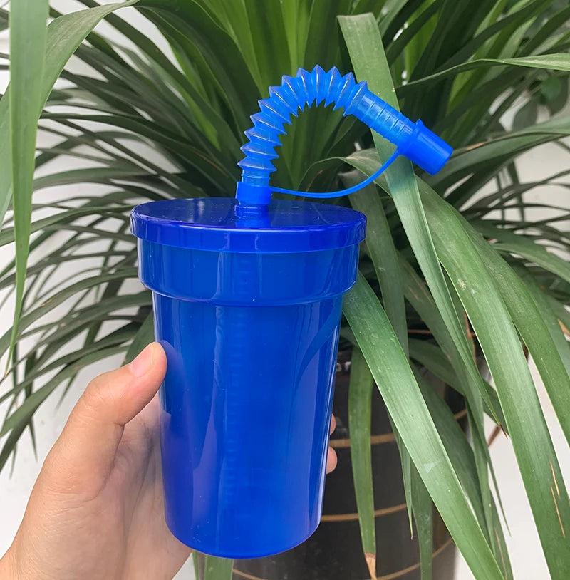 A hand holds a bright blue reusable sipper cup with a blue straw and lid, set against a background of green foliage.