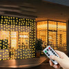 A hand holds a remote control for USB-powered fairy curtain string lights illuminating a storefront window.