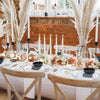 A rustic dining table setting featuring a centerpiece of dried foliage and candles against a brick fireplace backdrop.