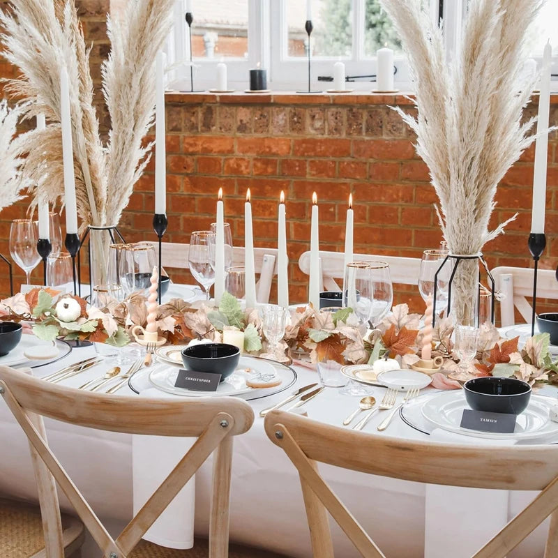 A rustic dining table setting featuring a centerpiece of dried foliage and candles against a brick fireplace backdrop.