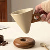 A hand holds a light beige ceramic coffee mug with a wooden base above a round wooden coaster.