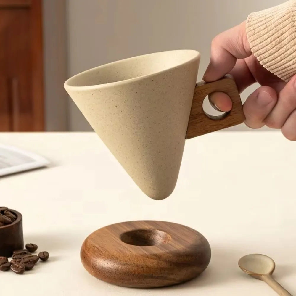 A hand holds a light beige ceramic coffee mug with a wooden base above a round wooden coaster.