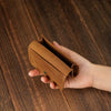 A hand holds a brown vintage leather card holder and cable organizer on a dark wooden surface.