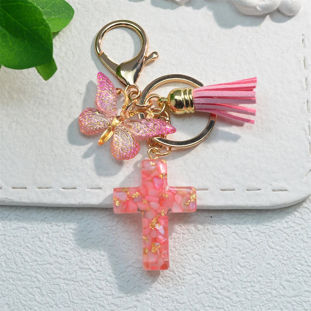 A gold-toned keychain featuring a pink glitter butterfly charm and a pink resin cross charm.