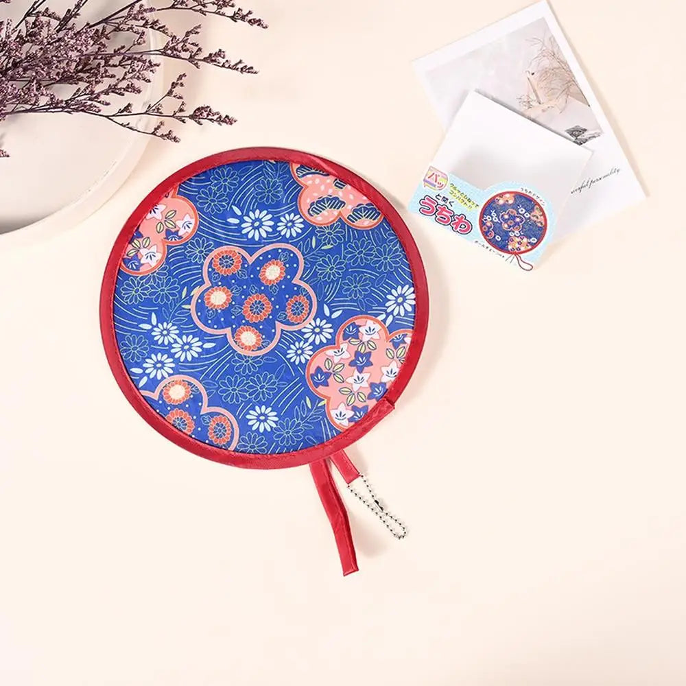 A round, fabric-covered handheld fan with blue and pink floral patterns rests on a light surface next to some paper.