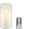 A white pillar candle in a glass holder with a visible flame, next to a black remote control.