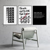 Three black and white motivational quote prints on a gray wall above a light wooden chair.