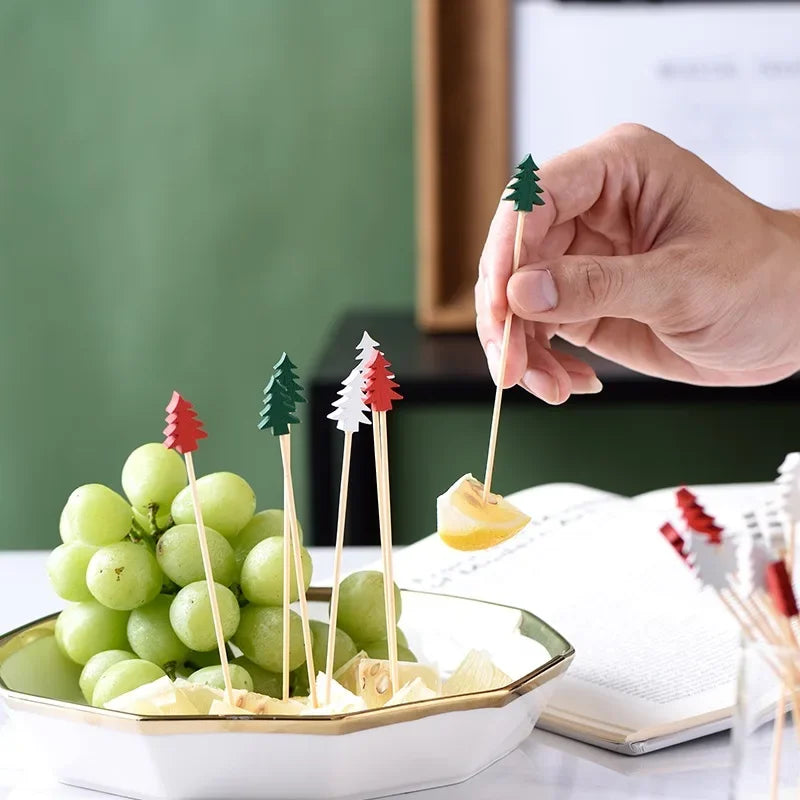 A hand is holding a green Christmas tree stick with a red accent, placed next to a bunch of green grapes on a white plate.