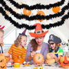 A woman and children are decorating a table with an orange pumpkin, surrounded by black tinsel garlands and Halloween-themed 
