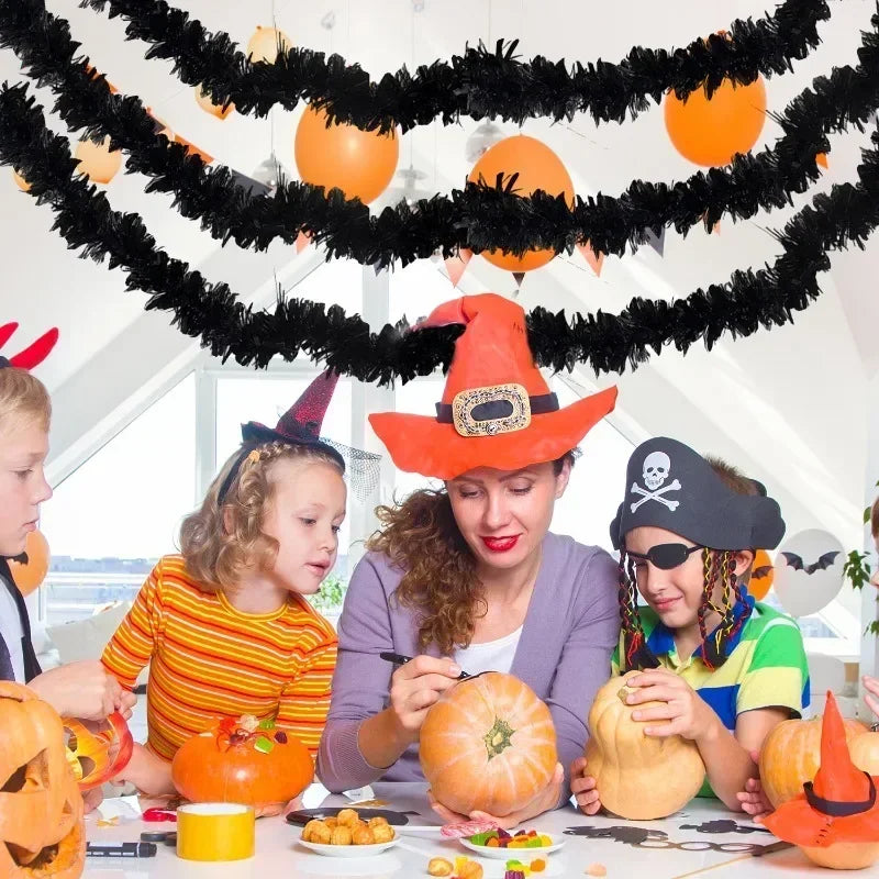 A woman and children are decorating a table with an orange pumpkin, surrounded by black tinsel garlands and Halloween-themed 