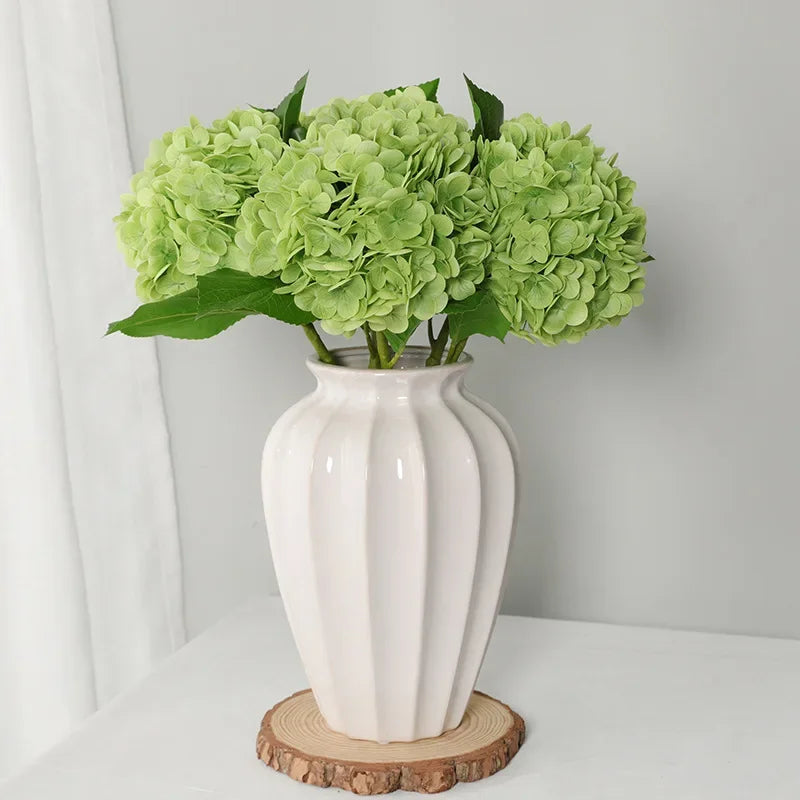A vase filled with light green artificial hydrangea flowers sits on a wooden coaster.